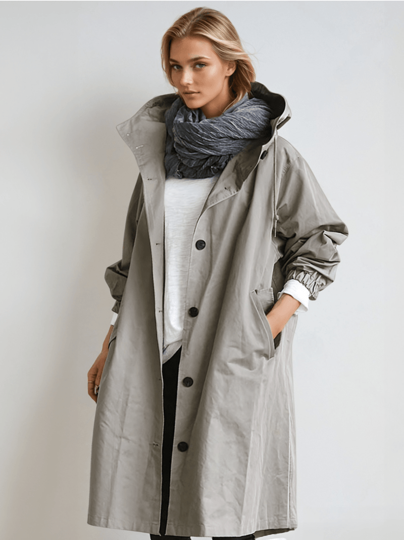 Model draagt de grijze trenchcoat met capuchon open over een wit shirt en donkere sjaal, met een hand in de zak. 1