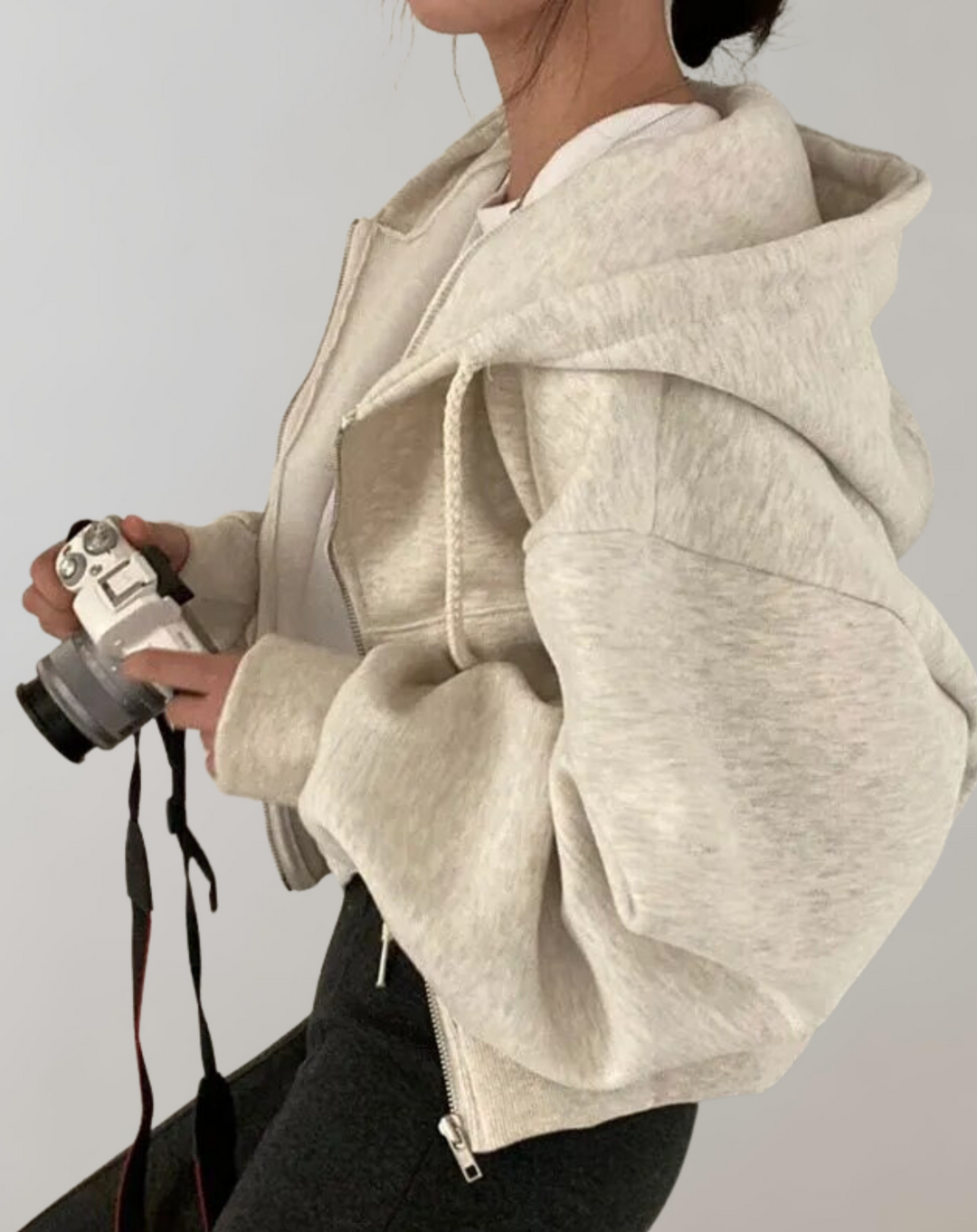 Zijaanzicht van de lichtgrijsbeige hoodie met wijde mouwen en capuchon, gedragen door een model dat een camera vasthoudt. 2