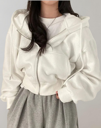 Model draagt de witte hoodie half open over een wit shirt en een grijze broek, met vlechtkoorden en zilverkleurige rits. 1