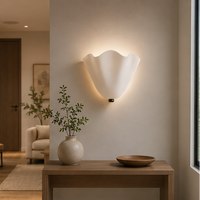 Verlichte witte wandlamp met golvende rand boven een houten tafel met vaas en schaal, met een woonkamer op de achtergrond. 8