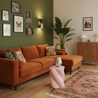 Wandlamp in donkergroen en crème tegen een olijfgroene muur, boven een oranje fluwelen hoekbank met roze bijzettafel. 9
