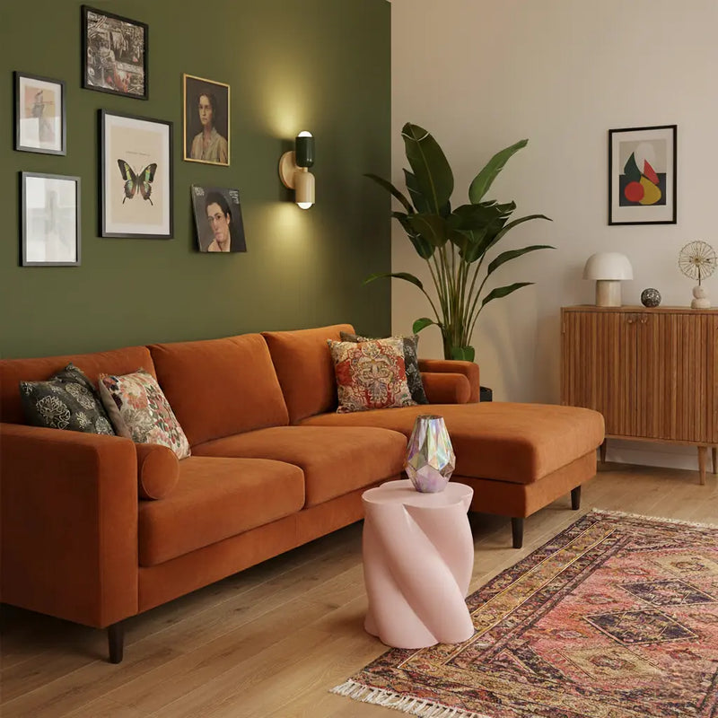 Wandlamp in donkergroen en crème tegen een olijfgroene muur, boven een oranje fluwelen hoekbank met roze bijzettafel. 9