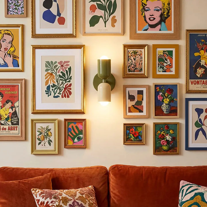 Brandende olijfgroene en crème wandlamp midden tussen een wand vol ingelijste posters, boven een oranje fluwelen bank met kussens. 6