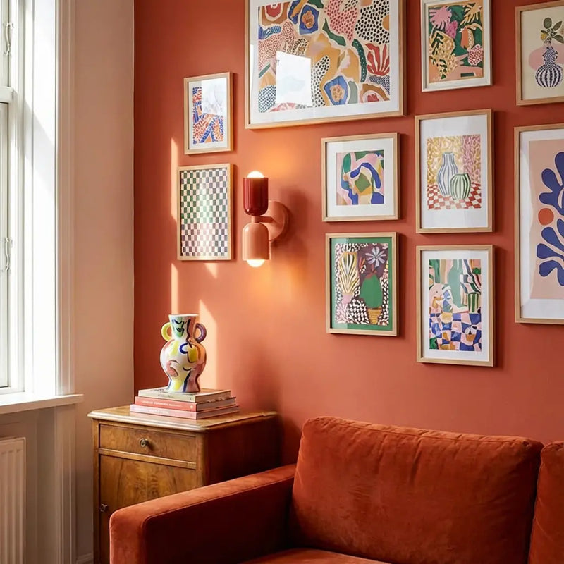 Wandlamp in rood en perzik tussen kleurrijke ingelijste kunstwerken op een terracotta muur, boven een oranje fluwelen bank. 5