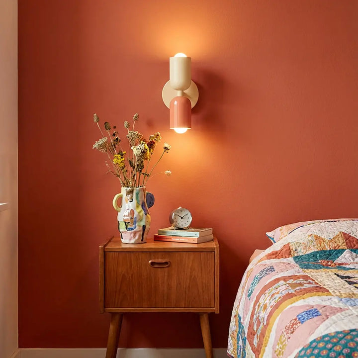 Brandende wandlamp in crème en zalmroze boven een vintage houten nachtkastje tegen een terracotta muur, naast een quilt op bed. 1