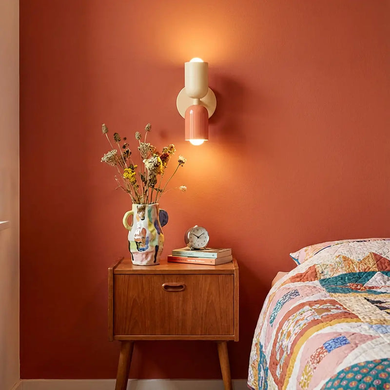 Brandende wandlamp in crème en zalmroze boven een vintage houten nachtkastje tegen een terracotta muur, naast een quilt op bed. 1