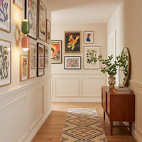 Groen-roze wandlamp aan een gang vol ingelijste posters, met een loper, houten dressoir en ronde spiegel. 3