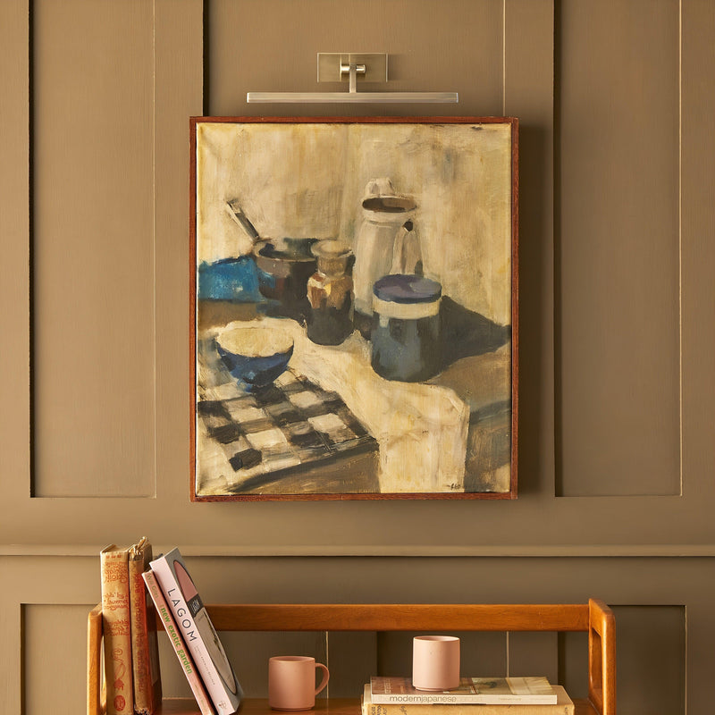 Uitgeschakelde messing schilderijlamp boven een ingelijst stilleven met kannen en een kom, in een kamer met houten 4