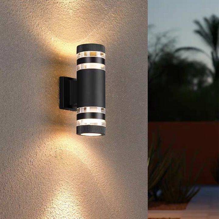 Zwarte cilindrische wandlamp met twee lichtbanden boven en onder, aan een ruwe muur met palmen op de achtergrond 5