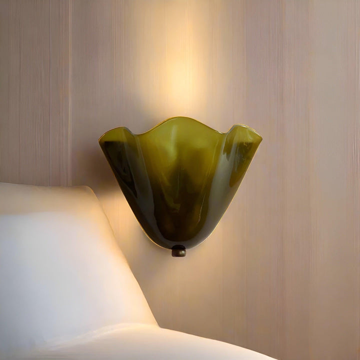 Close-up van de verlichte olijfgroene glazen wandlamp met golvende rand, naast een wit kussen, met warm licht langs de muur. 2