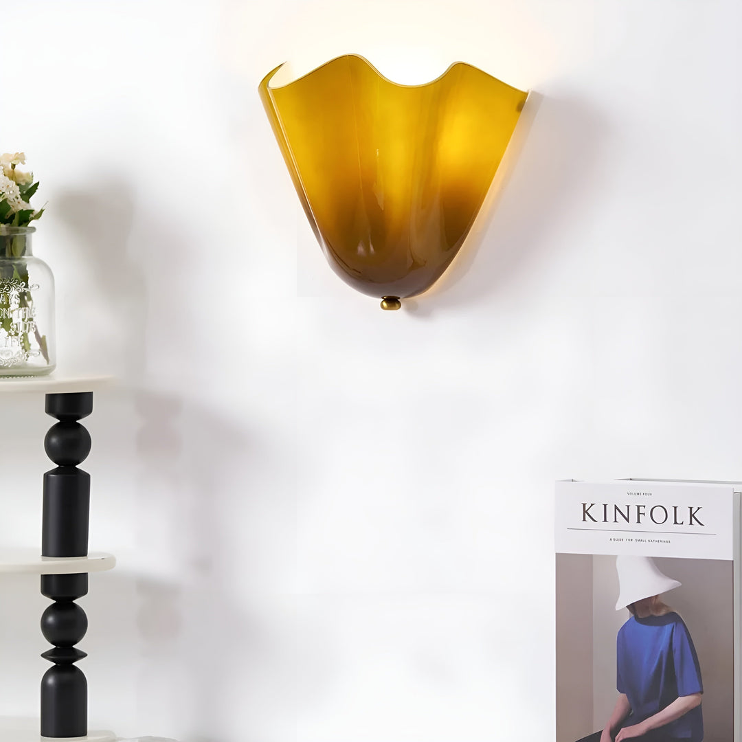Verlichte geelbruine glazen wandlamp op een witte muur, naast een zwarte houten kandelaar en een glazen vaas met bloemen. 5
