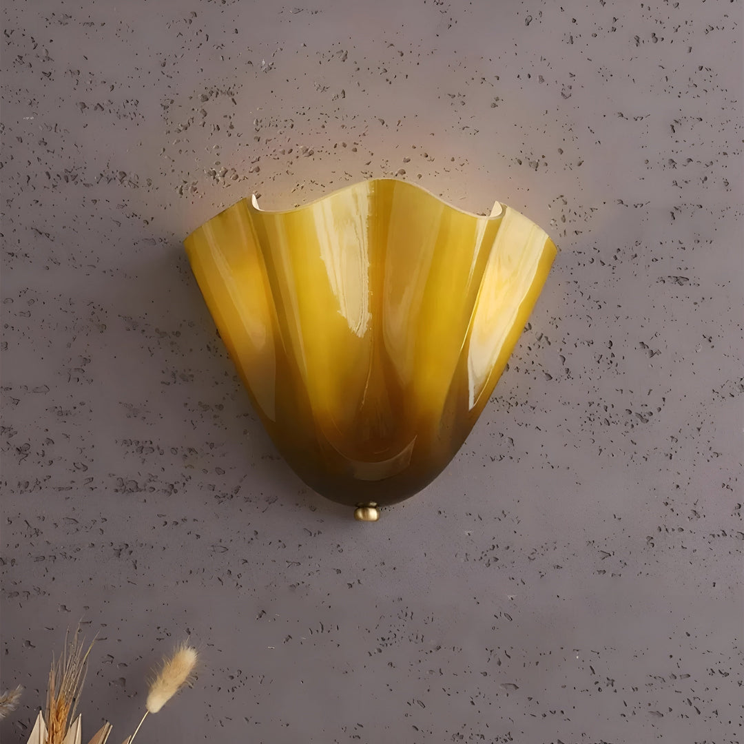 Close-up van de geelbruine glazen wandlamp met glanzende plooien, tegen een grijze structuurmuur met droge grassen onderin beeld. 11