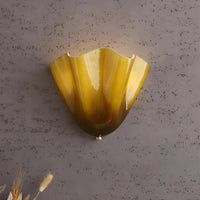 Close-up van de geelbruine glazen wandlamp met glanzende plooien, tegen een grijze structuurmuur met droge grassen onderin beeld. 11