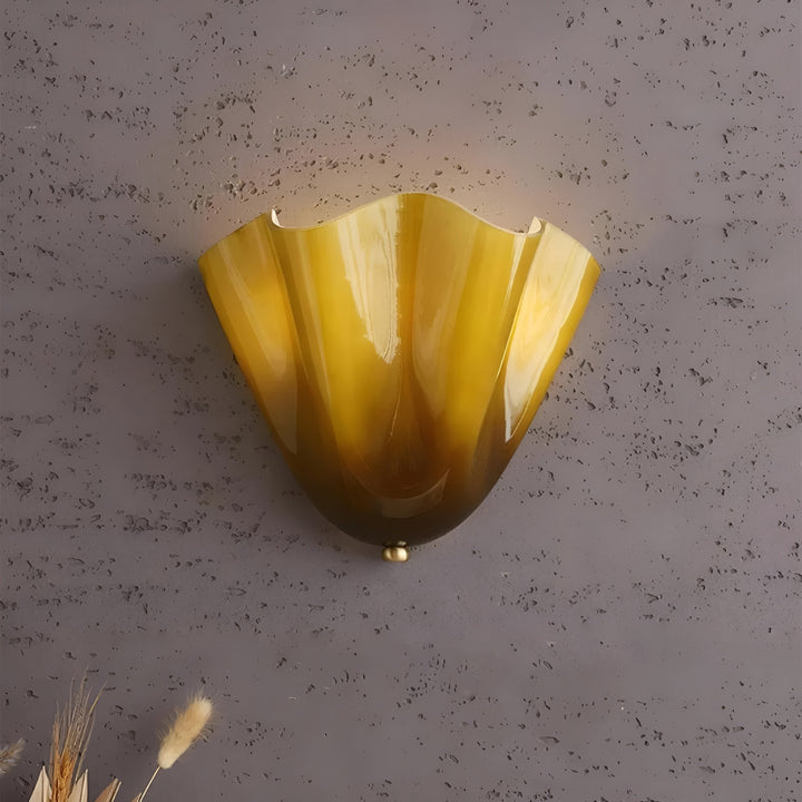 Close-up van de geelbruine glazen wandlamp met glanzende plooien, tegen een grijze structuurmuur met droge grassen onderin beeld. 11