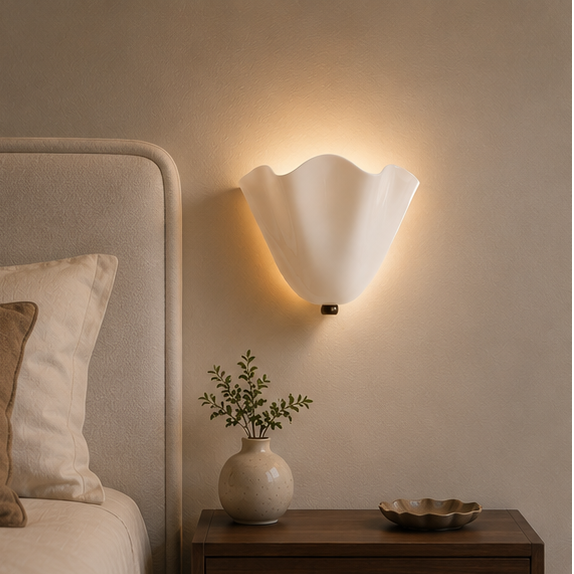 Verlichte witte wandlamp met golvende rand boven een nachtkastje met vaasje en schaaltje naast een bed in een slaapkamer. 1