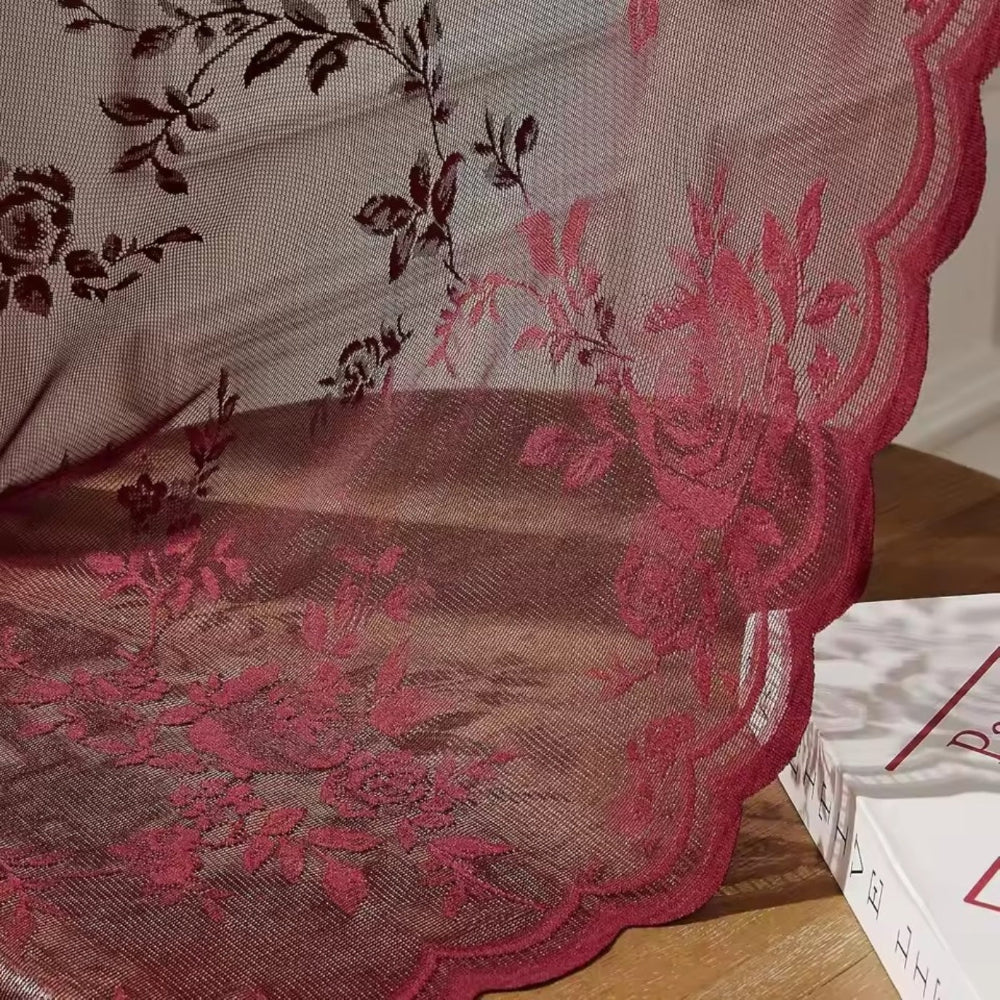 Close-up van rode kantstof met rozenmotief en golvende zoom, die over een houten tafel naast een boek valt. 3