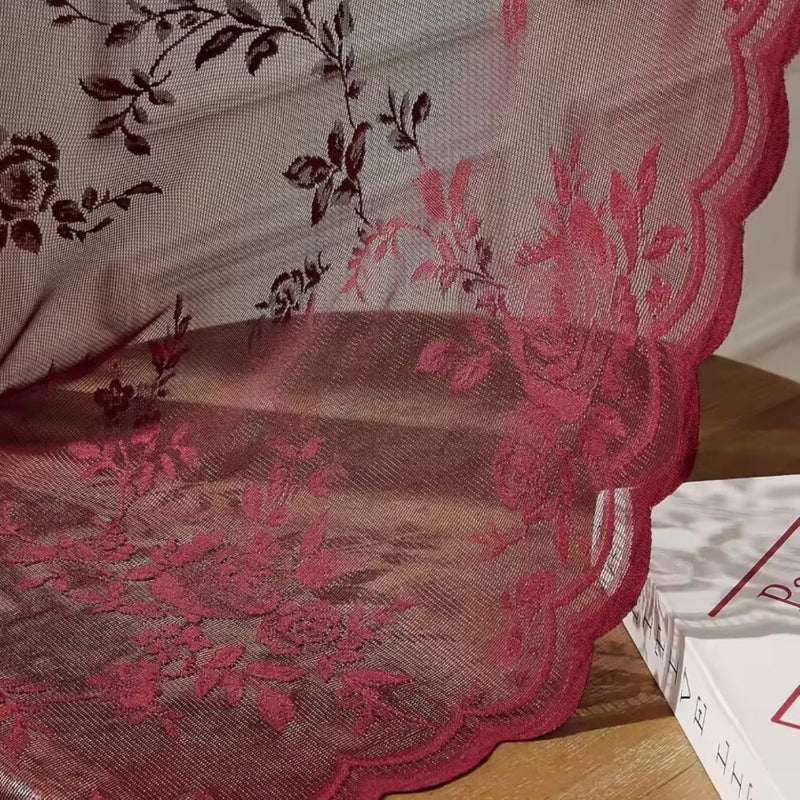 Close-up van rode kantstof met rozenmotief en golvende zoom, die over een houten tafel naast een boek valt. 3
