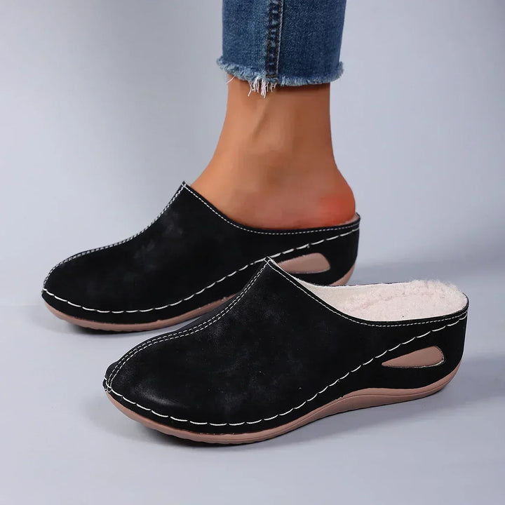 Zwarte pantoffels met fleecevoering gedragen door een model in jeans, zijaanzicht met ovale uitsparing in de wigzool. 2
