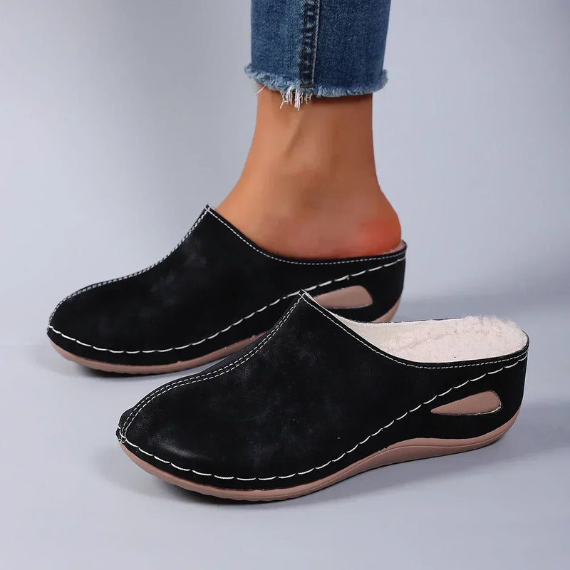 Zwarte pantoffels met fleecevoering gedragen door een model in jeans, zijaanzicht met ovale uitsparing in de wigzool. 2