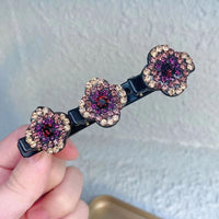 Close-up van een haarspeld met drie bloemen van strass in paarse, roze en goudkleurige tinten, vastgehouden in een hand. 2