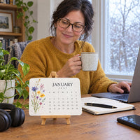 Kalenderkaart met wilde bloemen op een houten bureau, terwijl een vrouw met koffiemok achter haar laptop werkt 6