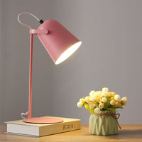 Roze versie van de bureaulamp met brandende kap, op een boek naast een groen vaasje met bloemen. 8