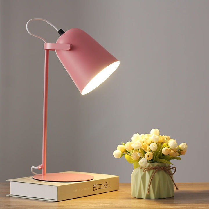 Roze versie van de bureaulamp met brandende kap, op een boek naast een groen vaasje met bloemen. 8