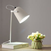 Witte versie van de bureaulamp met brandende kap, op een boek naast een groen vaasje met bloemen. 11