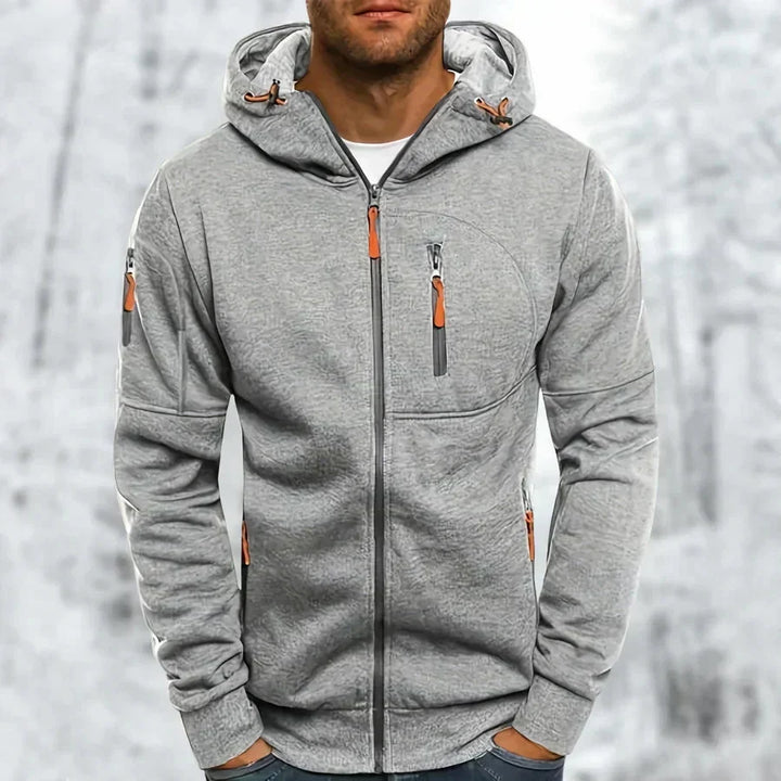 Man draagt de lichtgrijze gemêleerde hoodie met oranje ritstrekkers, geborduurde structuur zichtbaar, voor een besneeuwde achtergrond. 8