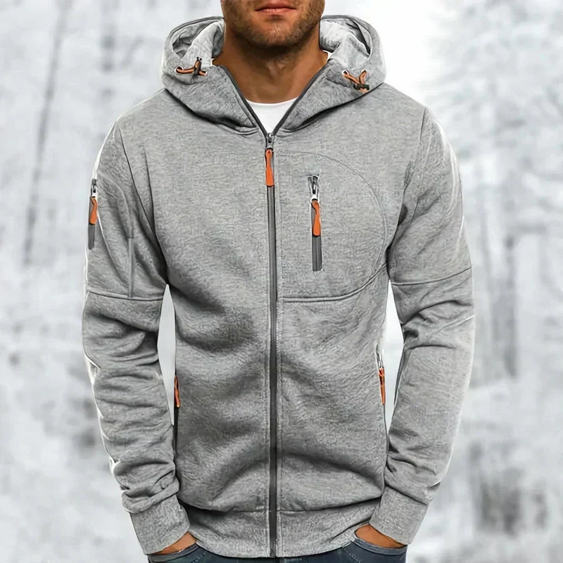 Man draagt de lichtgrijze gemêleerde hoodie met oranje ritstrekkers, geborduurde structuur zichtbaar, voor een besneeuwde achtergrond. 8