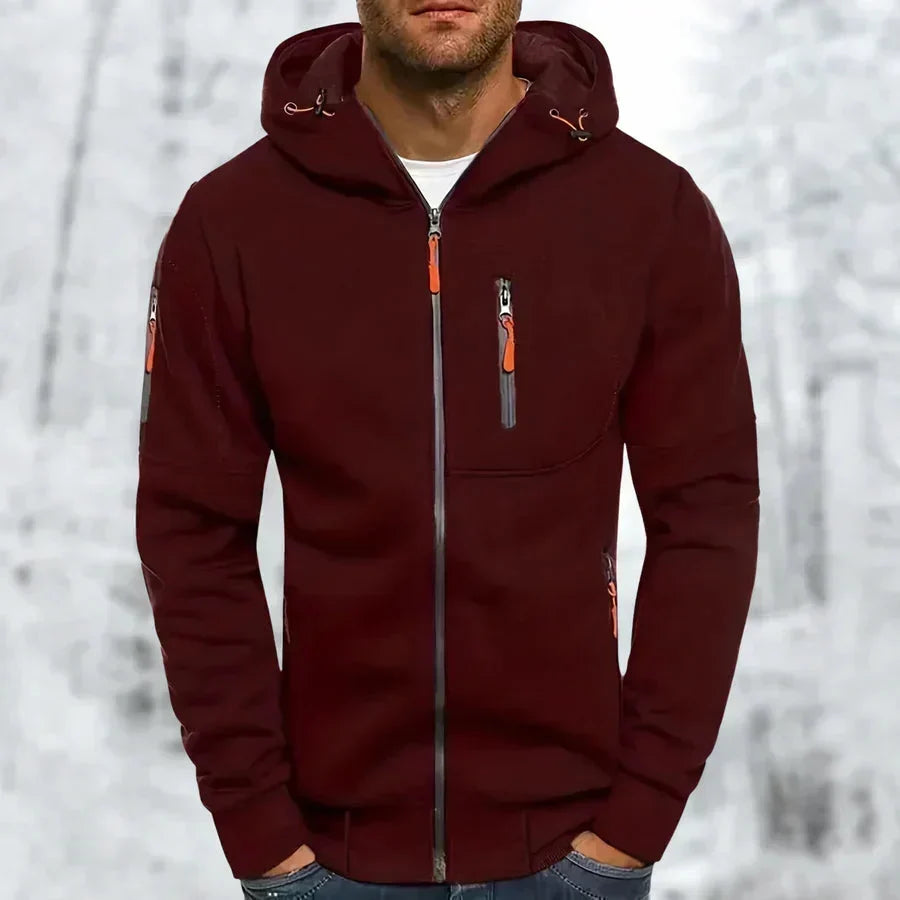 Man draagt de bordeauxrode hoodie met oranje ritstrekkers en grijze rits, handen in de zakken, met winters bos op de achtergrond. 4