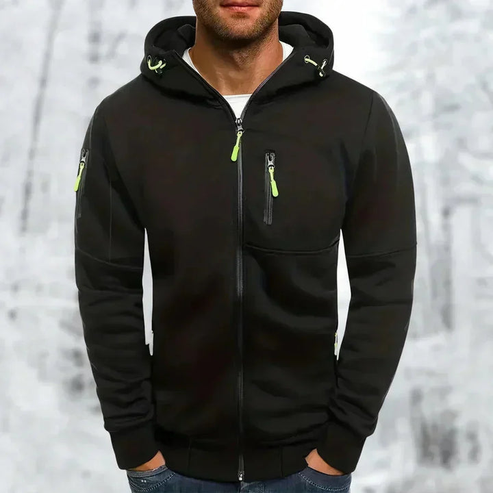 Zwarte hoodie met limoengroene ritstrekkers en capuchon, gedragen door een man met handen in de zakken voor een winterse achtergrond. 7