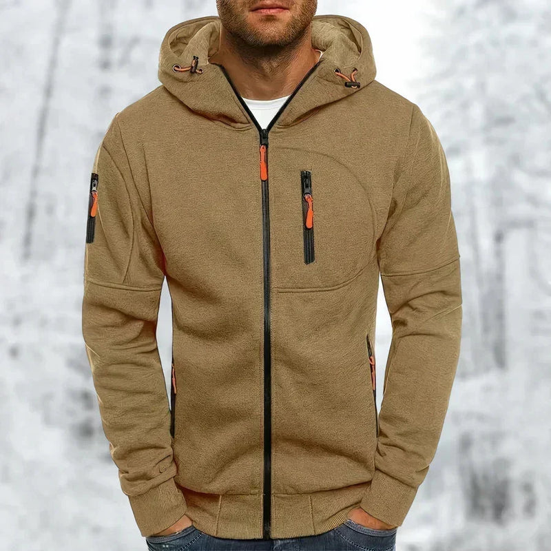 Man in de zandbruine hoodie met oranje ritstrekkers en zwarte rits, borstzakje en ribboorden zichtbaar, tegen een sneeuwachtergrond. 10