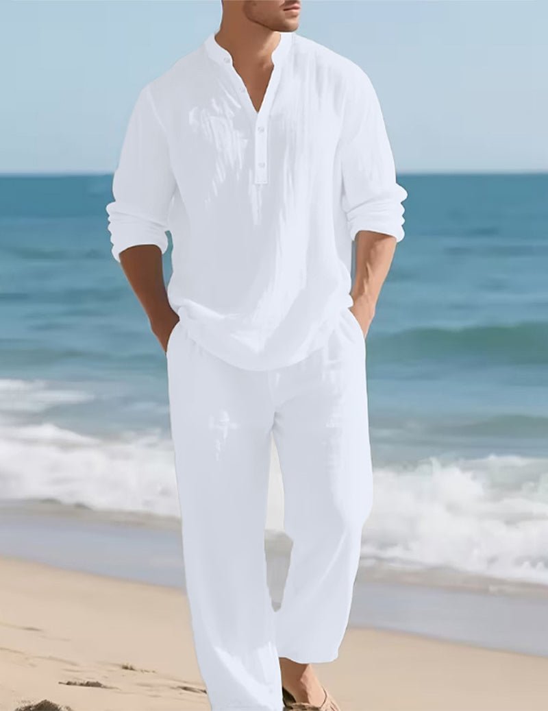 Model in de witte set loopt met handen in de zakken over het strand, zee en golven op de achtergrond. 2