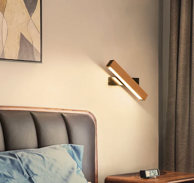 Houten wandlamp met zwarte steun in diagonale stand boven een nachtkastje met digitale wekker naast een gecapitonneerd hoofdbord. 8