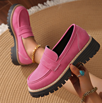 Felroze suèdelook loafers met zwarte profielzool, een model houdt één schoen vast op een rieten kleed met gouden armbanden. 18