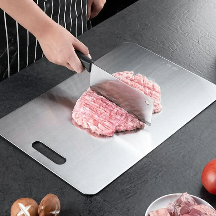 Metalen snijplank op een donker aanrecht waarop een hakmes gehakt vlees fijnhakt, met een uitgesneden handgreep in de plank. 1