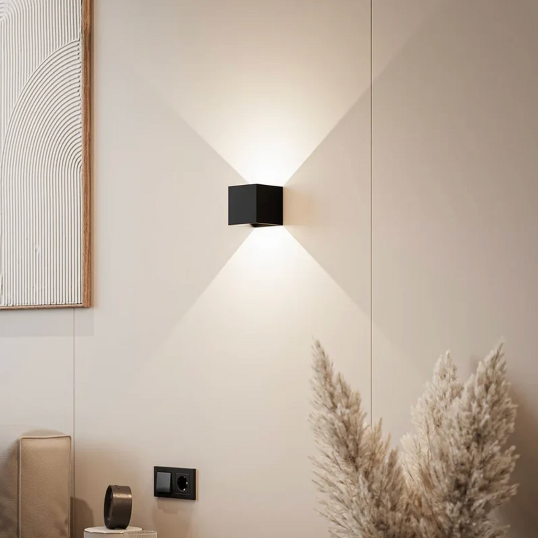 Zwarte kubusvormige wandlamp op een beige muur die licht naar boven en beneden werpt, naast een schilderij en pampasgras. 1