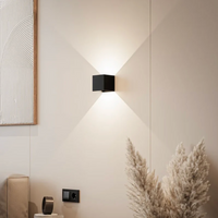 Zwarte kubusvormige wandlamp op een beige muur die licht naar boven en beneden werpt, naast een schilderij en pampasgras. 1
