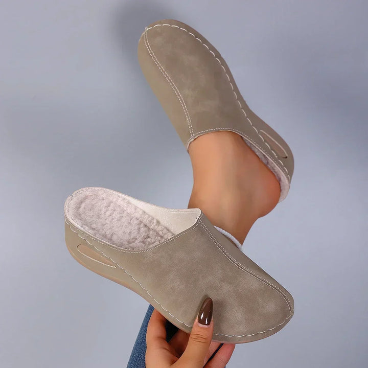 Beige pantoffels met zichtbare fleecevoering aan de binnenkant, één vastgehouden in de hand en één aan de voet. 6