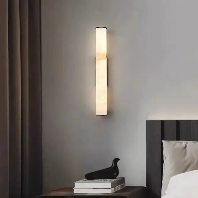 Slanke verticale marmeren wandlamp tussen gordijnen en een donker bed, met boeken en een zwart vogelbeeldje op het nachtkastje. 14