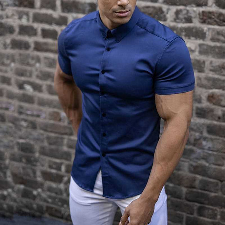 Blauw overhemd met korte mouwen, gedragen door een man in een witte broek voor een bakstenen muur. 3