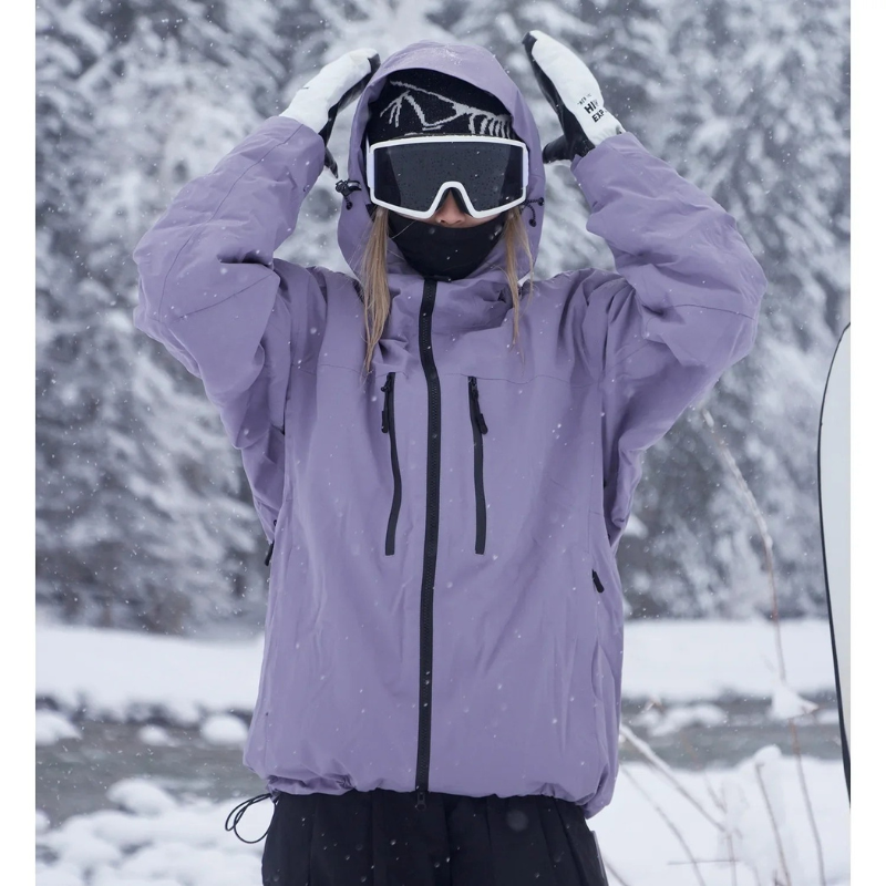Model in lila ski-jack met zwarte ritsen trekt de capuchon op met beide handen, in een besneeuwd bos. 1
