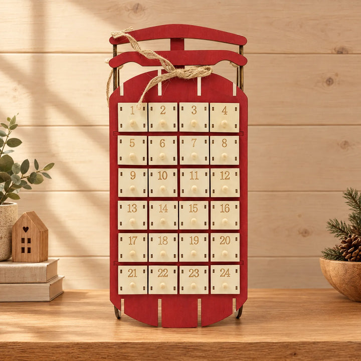 Rode houten sleekalender met 24 vakjes en jutetouw, staand op een houten plank met kerstdecoratie en een houten huisje ernaast. 1