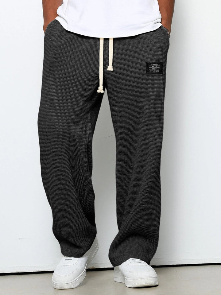 Voorkant van de wafelgebreide broek in donkergrijs, gedragen door een model met wit shirt, wijde pijpen en een crèmekleurig trekkoord. 1