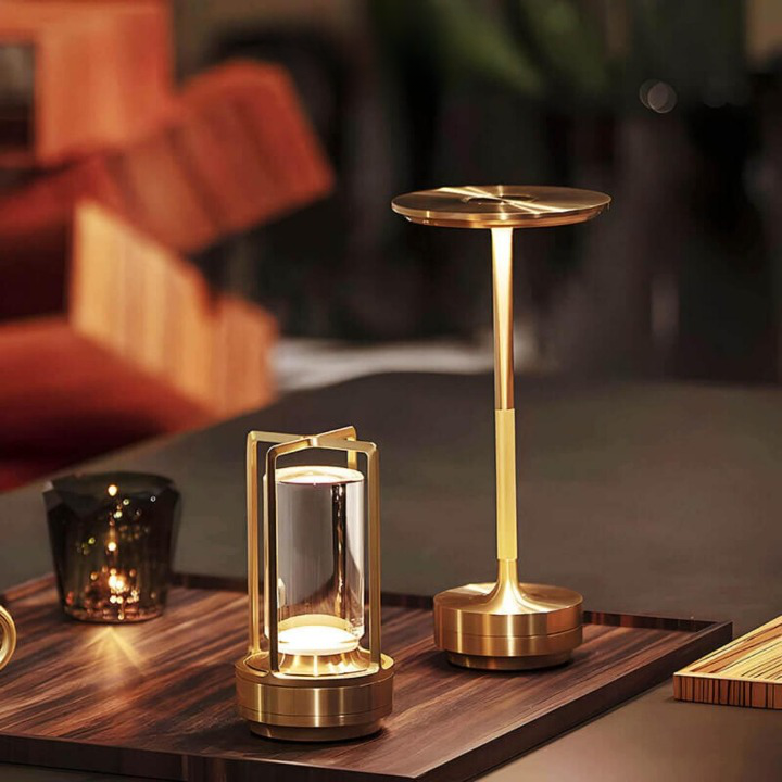 Goudkleurige lamp met open frame naast een hogere gouden paddenstoelvormige tafellamp op een houten dienblad. 7
