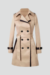 Beige dubbelrijige trenchcoat met donkere bies, ceintuur en bandjes aan de mouwen, los neergelegd op een lichte achtergrond. 1