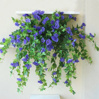 Paarsblauwe kunstbloemen met lange groene ranken die van een witte wandplank naar beneden vallen tegen een crèmekleurige muur. 4