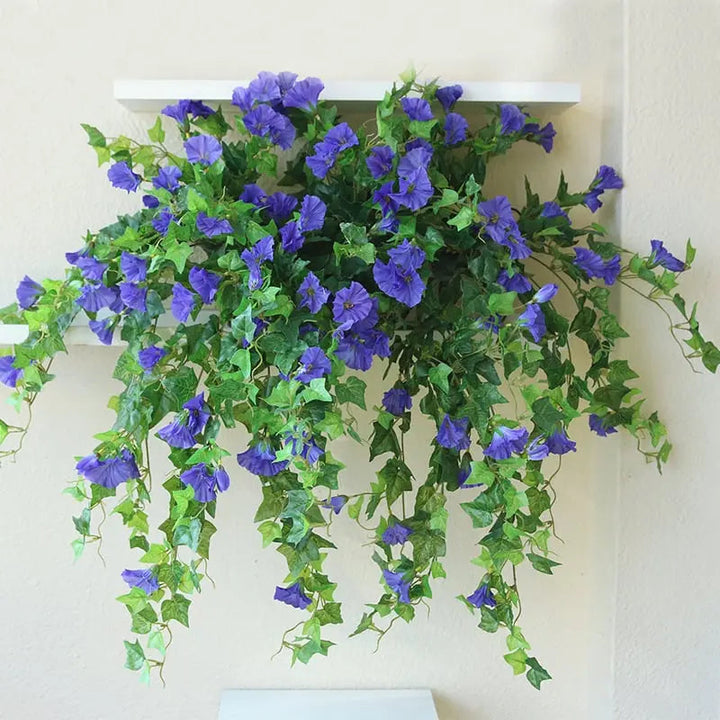 Paarsblauwe kunstbloemen met lange groene ranken die van een witte wandplank naar beneden vallen tegen een crèmekleurige muur. 4