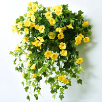 Dichte bos gele kunstbloemen met groene bladeren, tegen een witte wand gehangen, met ranken die naar beneden vallen. 5